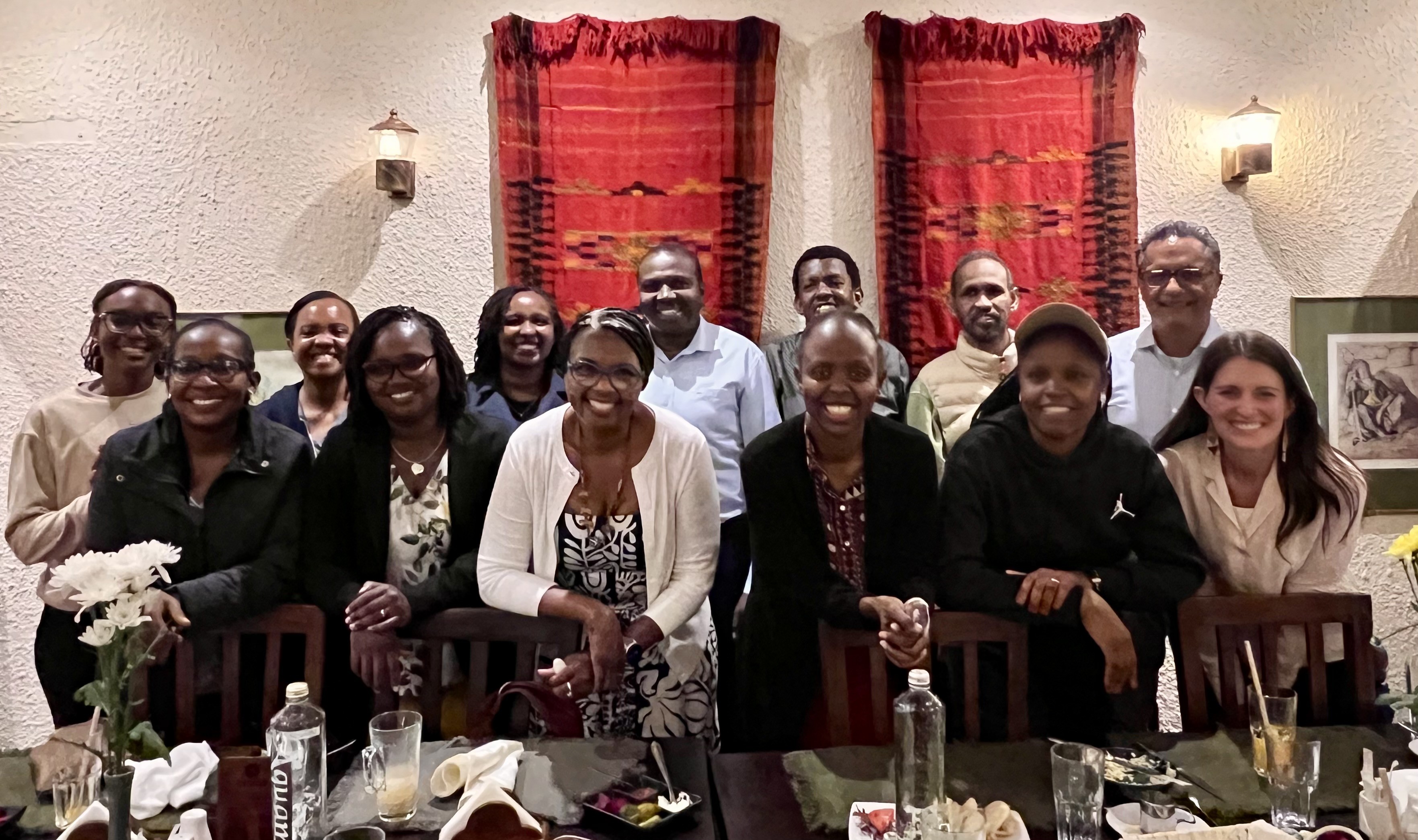 A group of people pose behind a dining table with orange tapestries in the background.