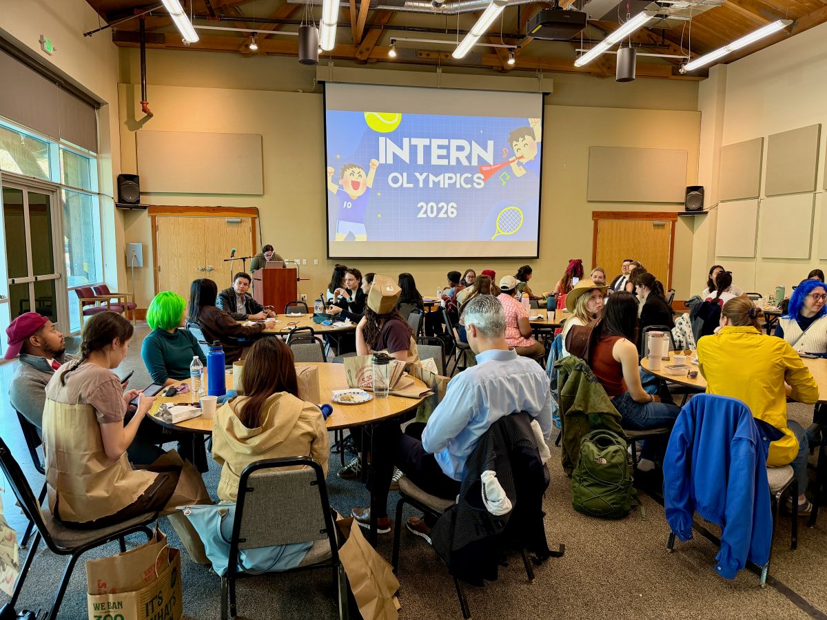 A group of people sit at tables viewing a slide that says "Intern Olympics 2026"