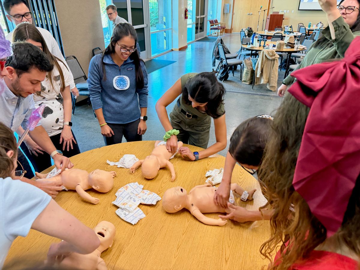 People stand around a table with infant dolls laid out for practice activities.