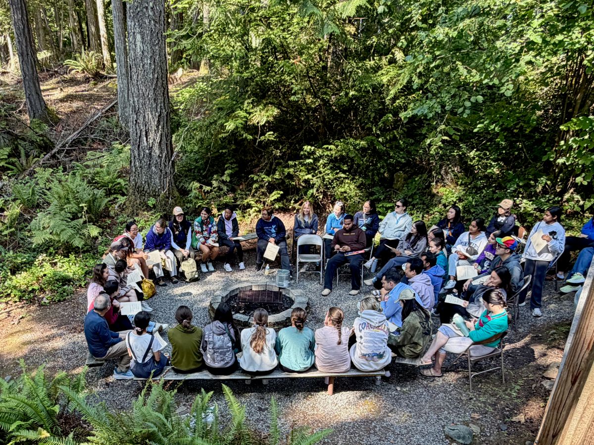 A view from above of a large group sitting outside in a circle in the woods. 