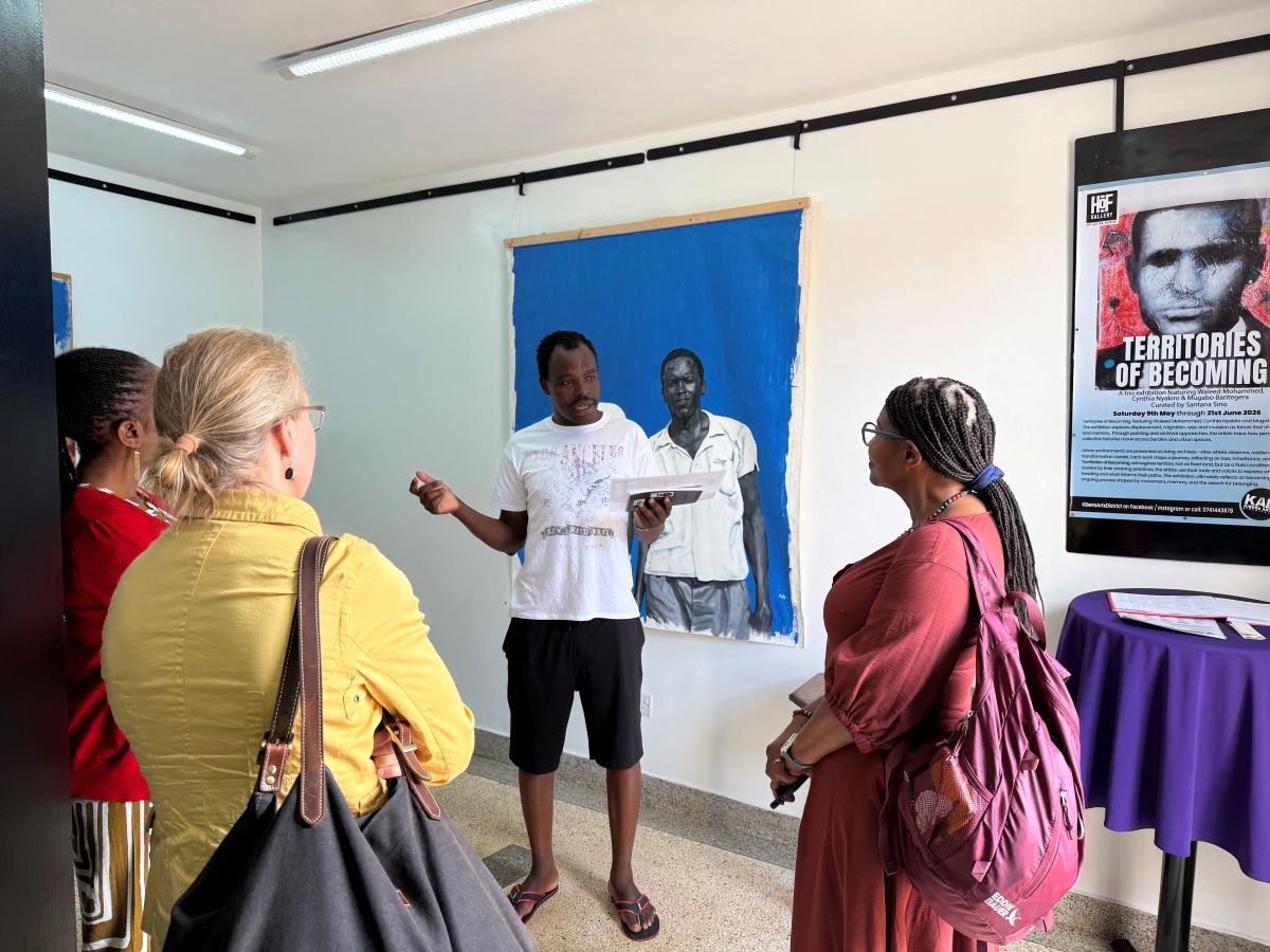 A person in a white shirt and black shorts speaks to a small group of people with a piece of artwork behind him depicting a figure on a bright blue background. 