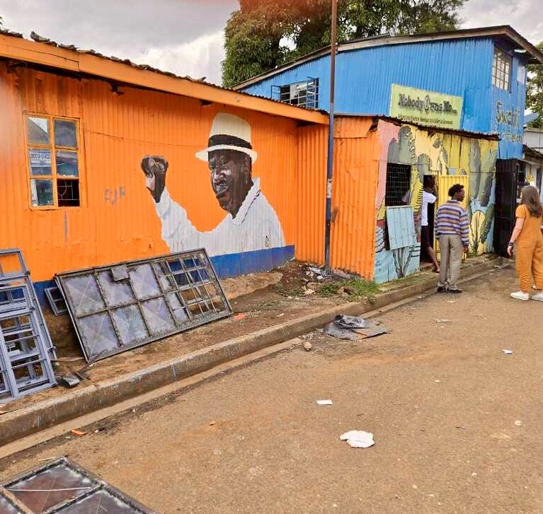 A mural painted on an orange metal wall shows a man in a hat holding up his right hand. 