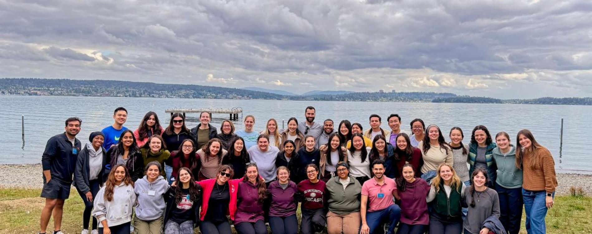 A large group of people pose for a group photo in front of a lake and cloudy sky. 