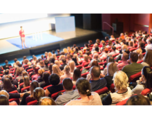 An image from the back of a lecture hall shows a large audience watching a speaker on stage. 